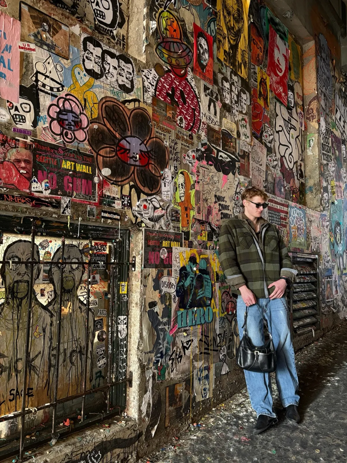 Zander Dent leaning against the Seattle art wall, a dense collage of stickers and posters